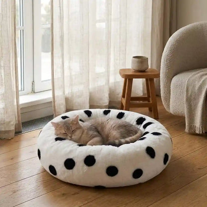 A fluffy round pet bed with black polka dots on a cream background, with a gray cat sleeping inside, set on a wooden floor in a bright room with curtains and a chair.